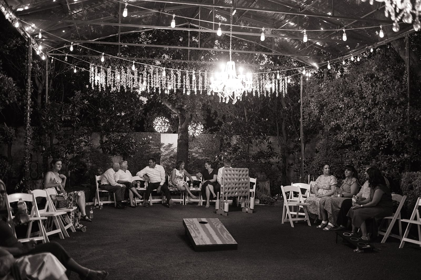 People stand and talk under string lights and a chandelier at an outdoor event with chairs arranged around a wooden archway at Magnolia Terrace in Frisco, Tx