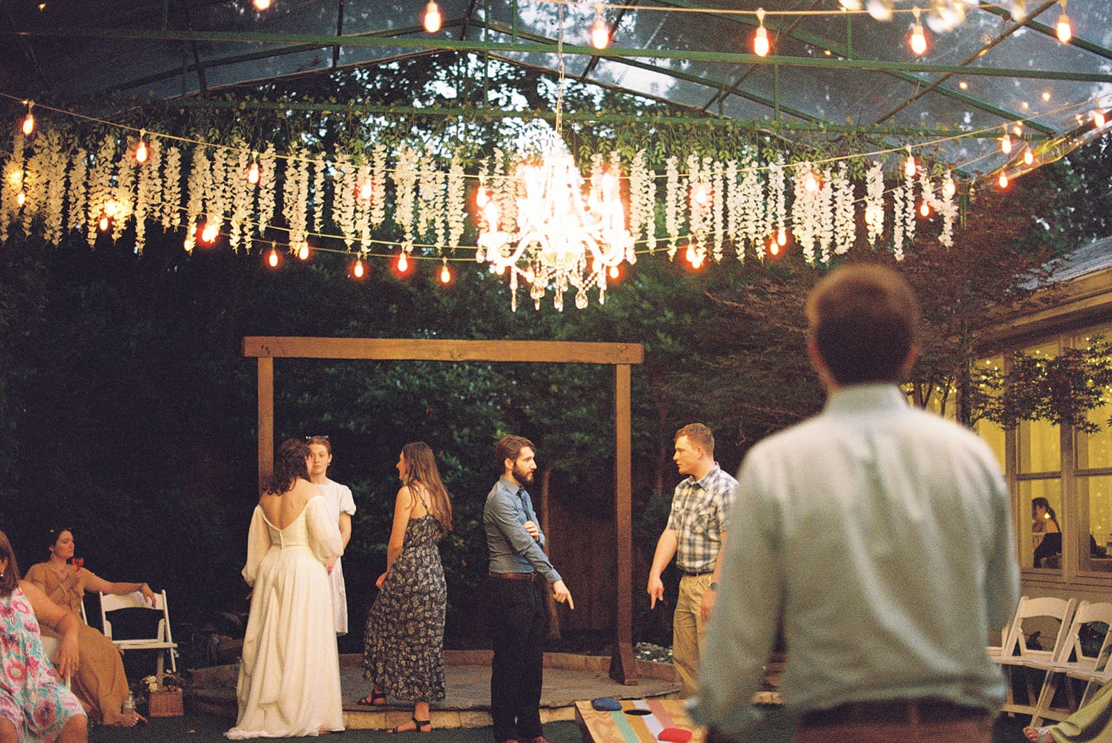 People stand and talk under string lights and a chandelier at an outdoor event with chairs arranged around a wooden archway.