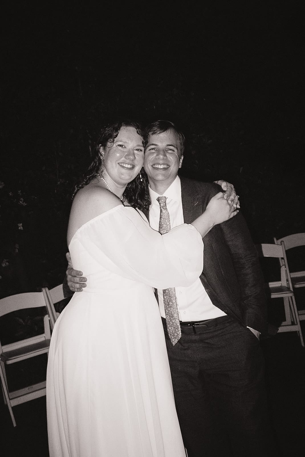 A couple in formal attire dances alone under chandeliers and string lights, surrounded by empty white chairs in an outdoor setting at night at Magnolia Terrace in Frisco, Tx