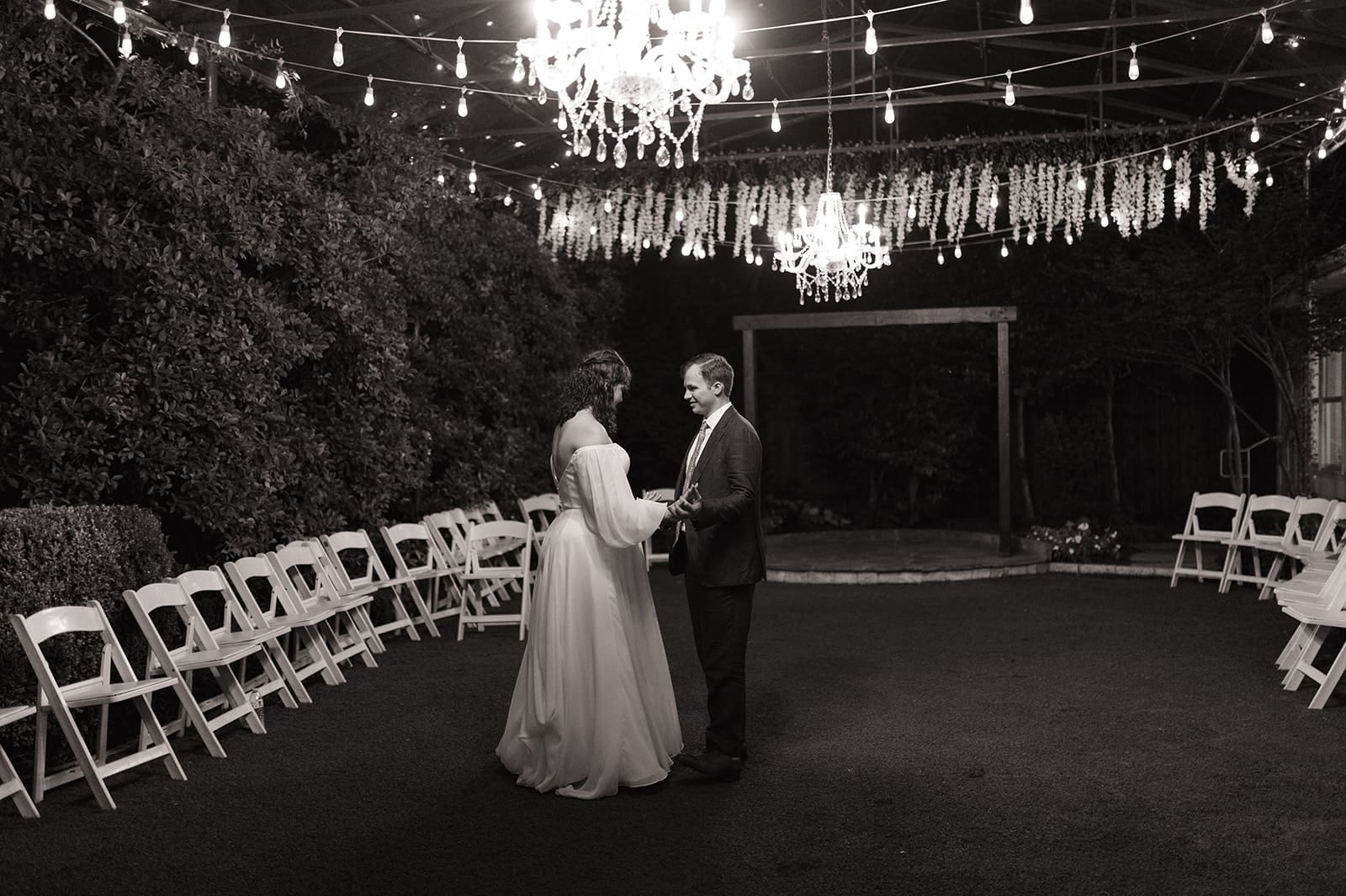 A couple in formal attire dances alone under chandeliers and string lights, surrounded by empty white chairs in an outdoor setting at night at Magnolia Terrace in Frisco, Tx