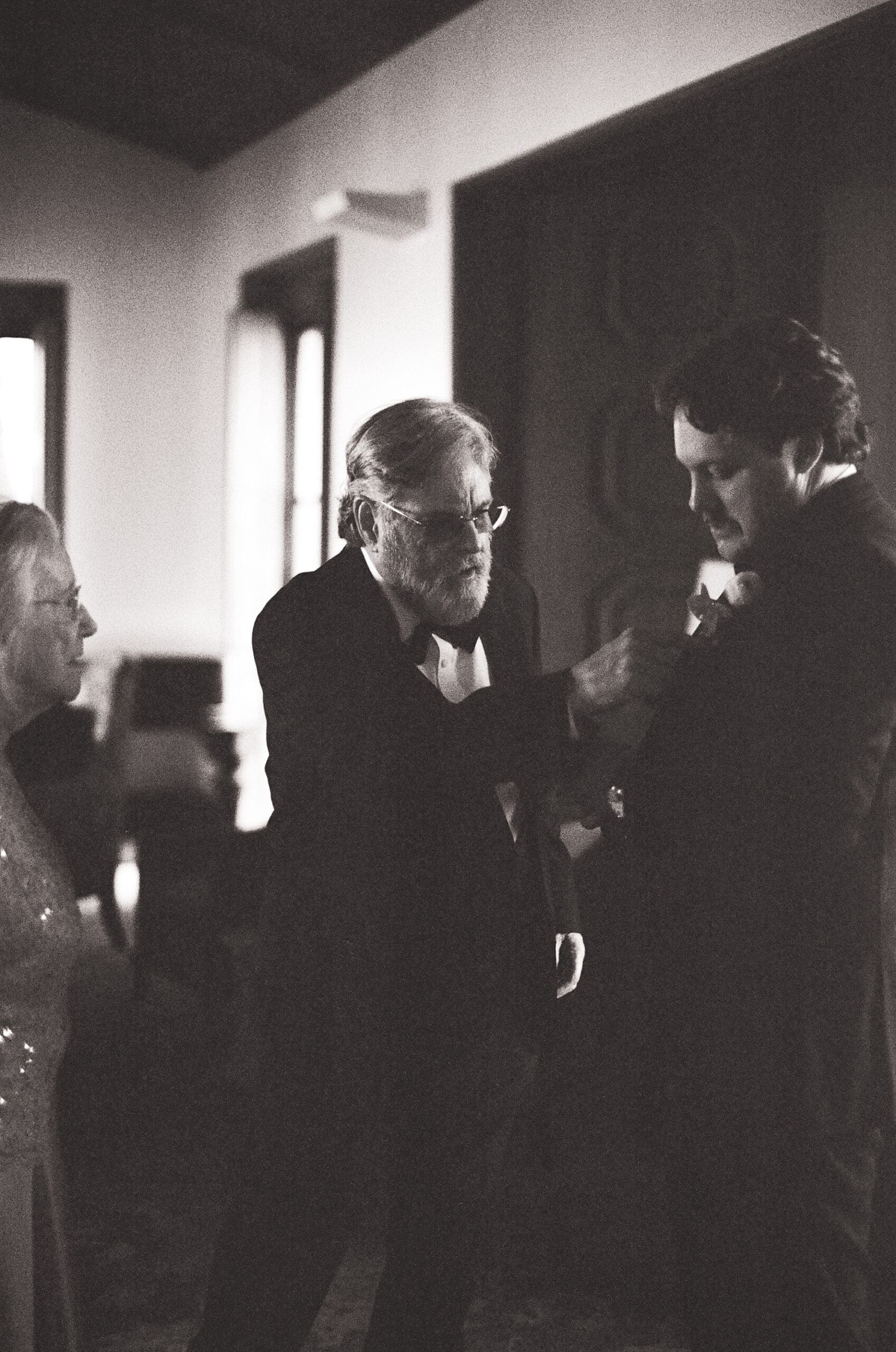 An older man adjusts a younger man's boutonniere in a dimly lit room, while another person stands nearby.