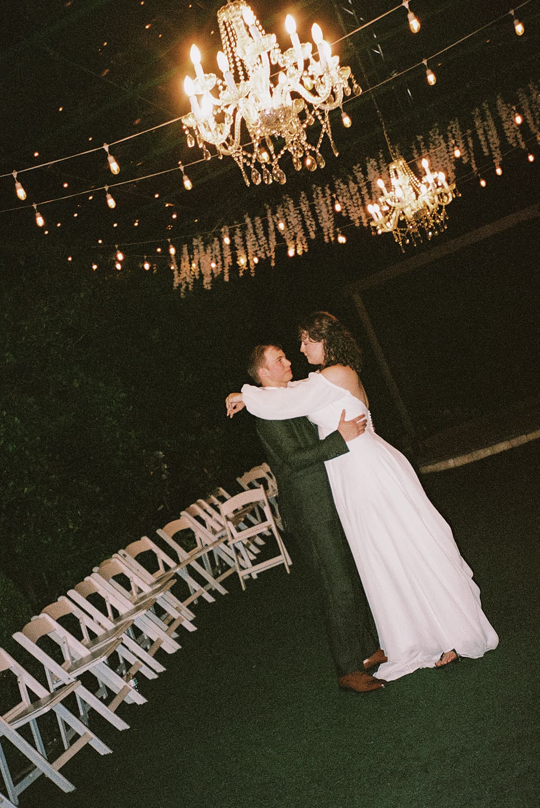 A couple in formal attire dances alone under chandeliers and string lights, surrounded by empty white chairs in an outdoor setting at night at Magnolia Terrace in Frisco, Tx