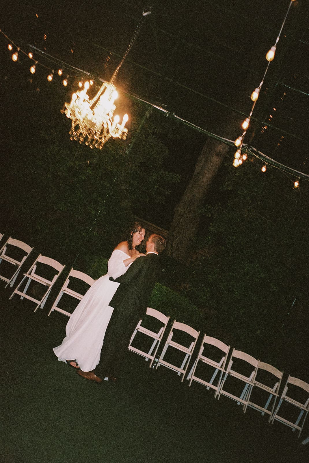 A couple in formal attire dances alone under chandeliers and string lights, surrounded by empty white chairs in an outdoor setting at night at Magnolia Terrace in Frisco, Tx
