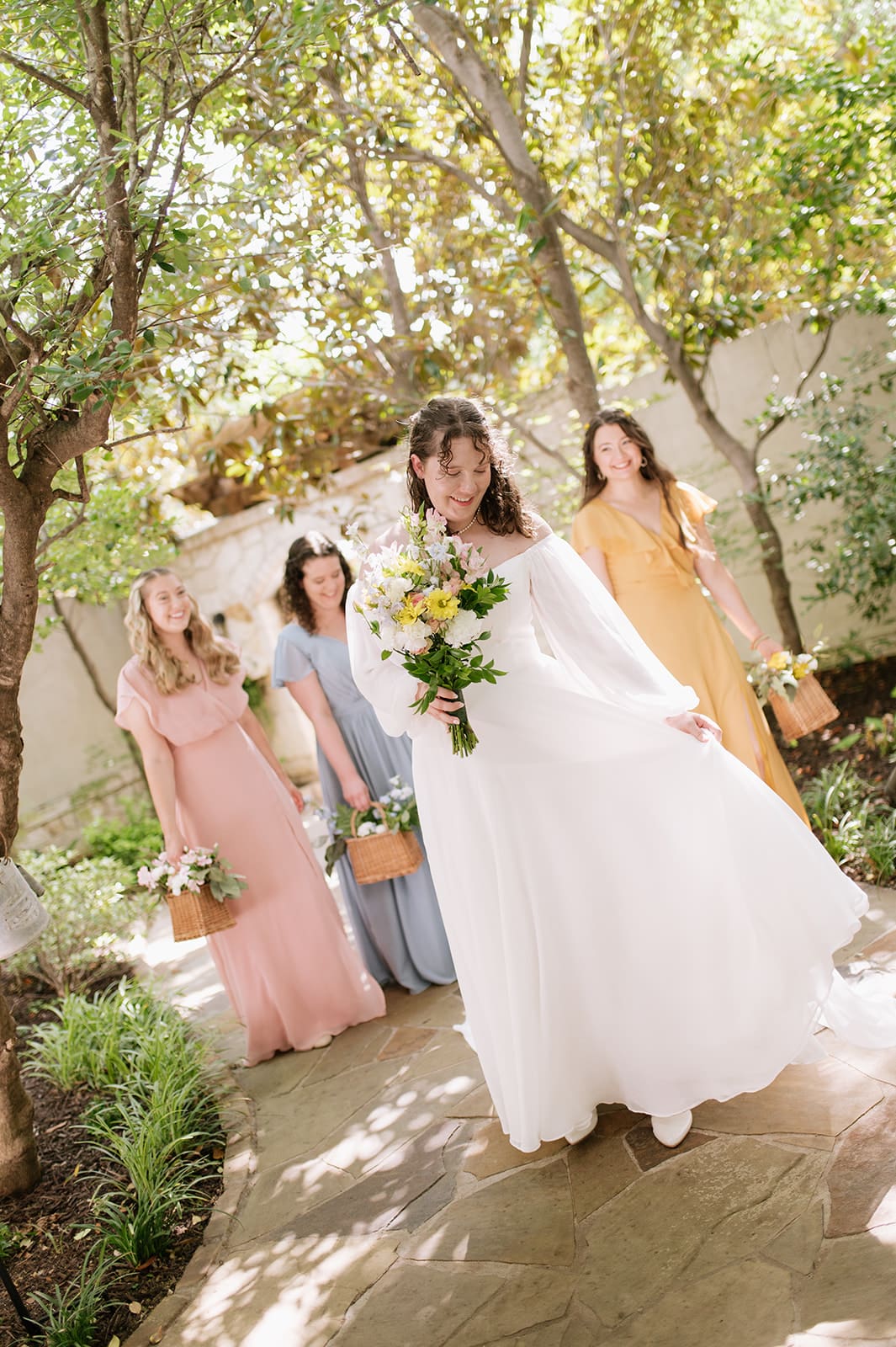 Four women in colorful dresses stand outdoors, smiling and laughing. The woman in white holds a bouquet, suggesting she is the bride, while the others hold baskets of flowers.