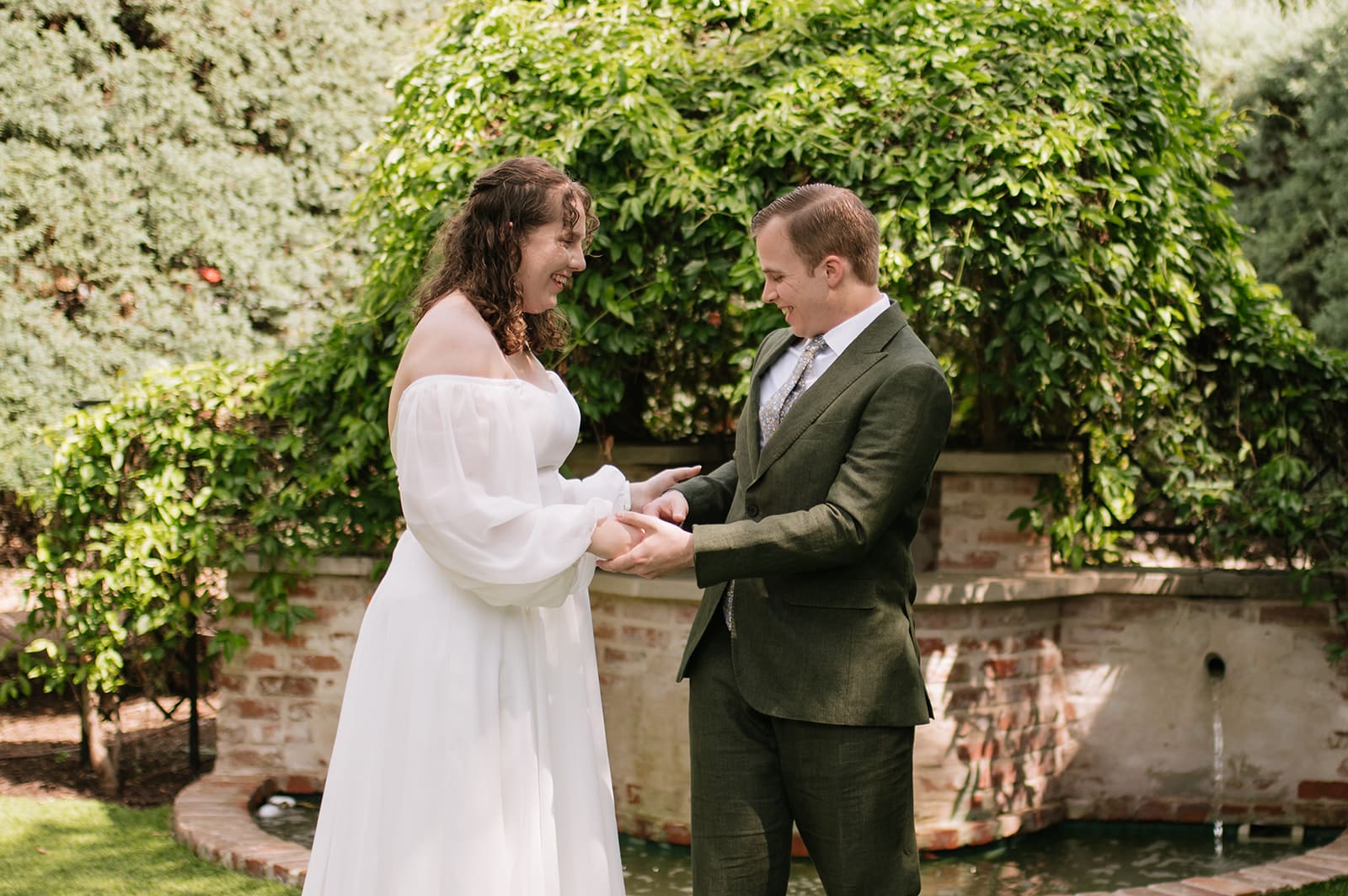 A bride in a white dress and a groom in a dark suit hold hands and smile at each other outdoors in front of greenery and a small fountain for a wedding at Magnolia Terrace in Frisco Tx