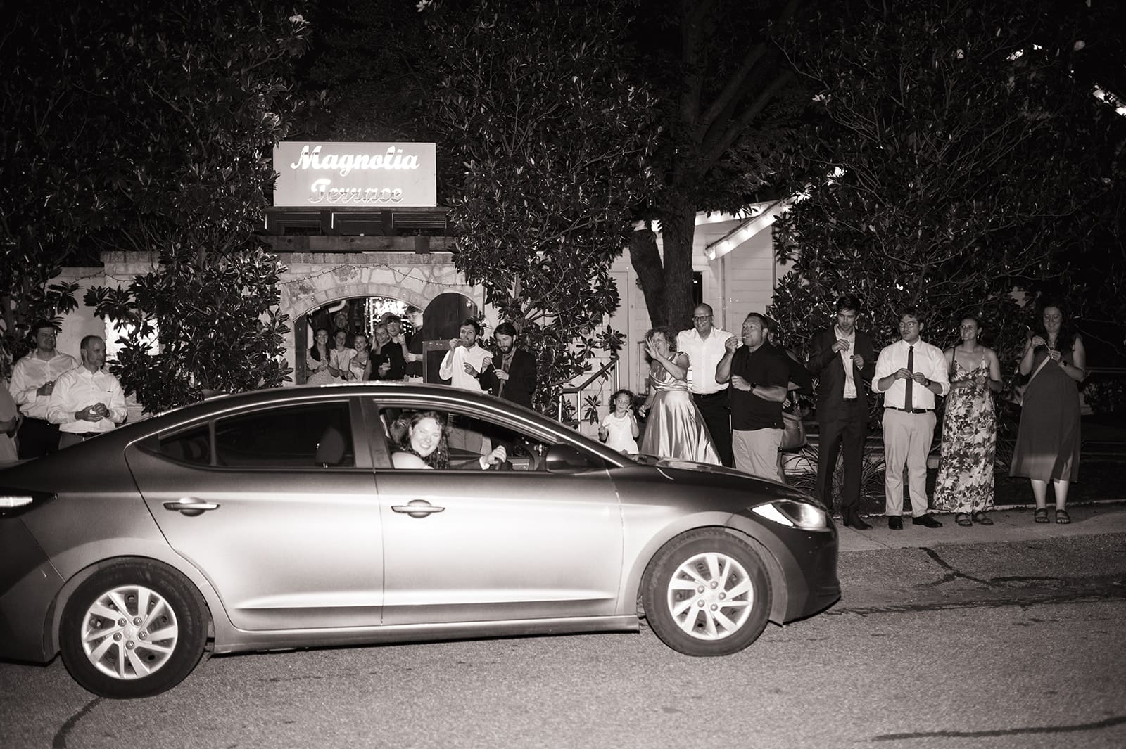 A car drives past a group of people standing on a sidewalk at night; some are dressed formally and appear to be watching or celebrating.