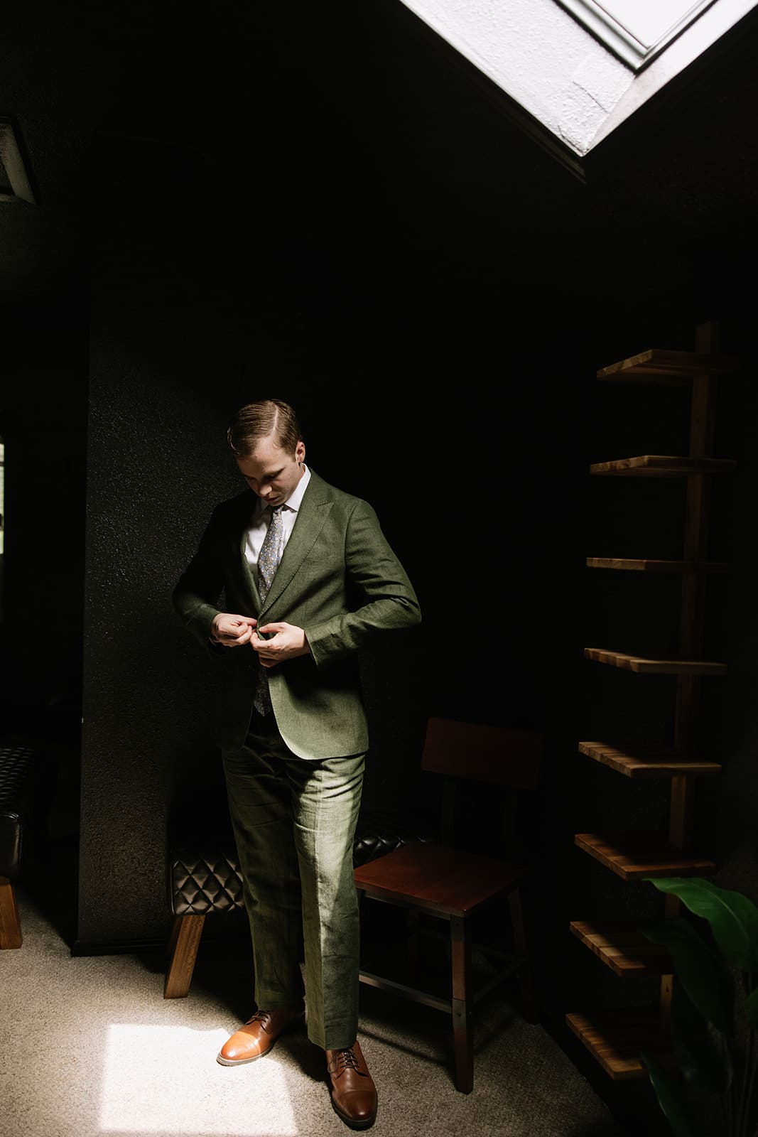 A man in a green suit stands indoors under a skylight, buttoning his jacket.