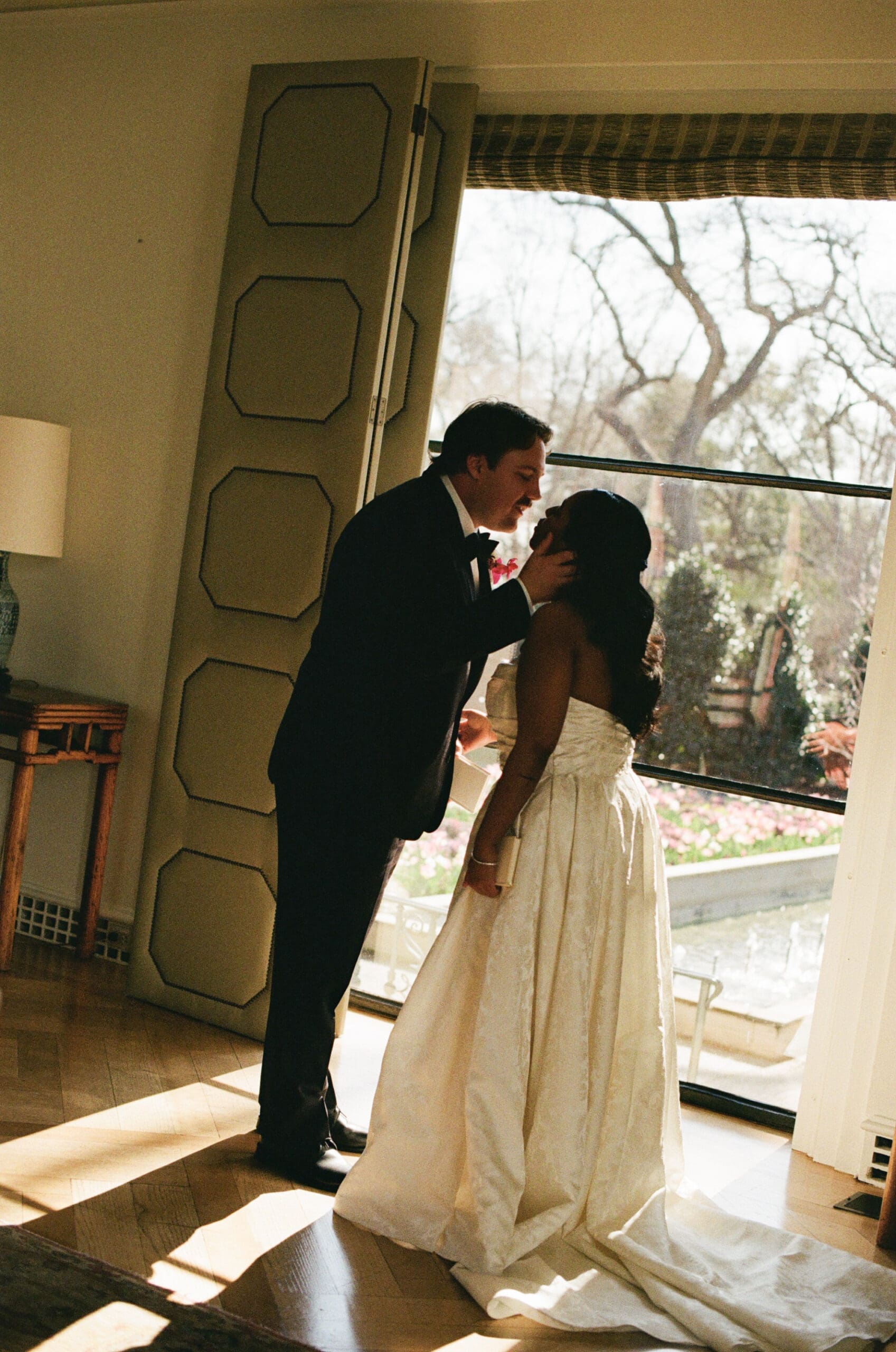 A woman in a white dress bends over inside a large window, reaching toward a man standing next to her for a Dallas Arboretum Wedding