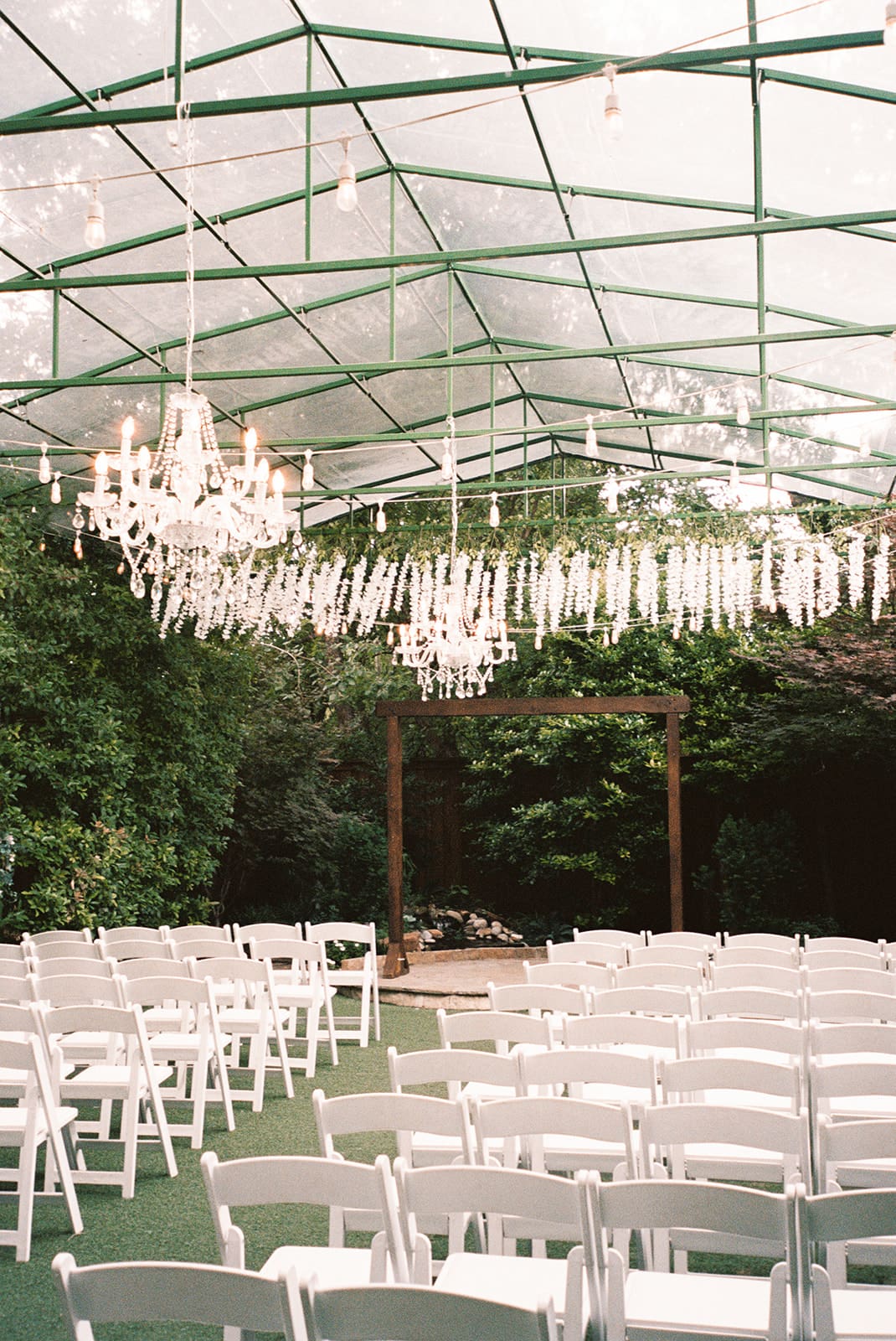 Rows of white chairs are arranged under a transparent tent with chandeliers and hanging lights, facing a wooden arch surrounded by greenery.