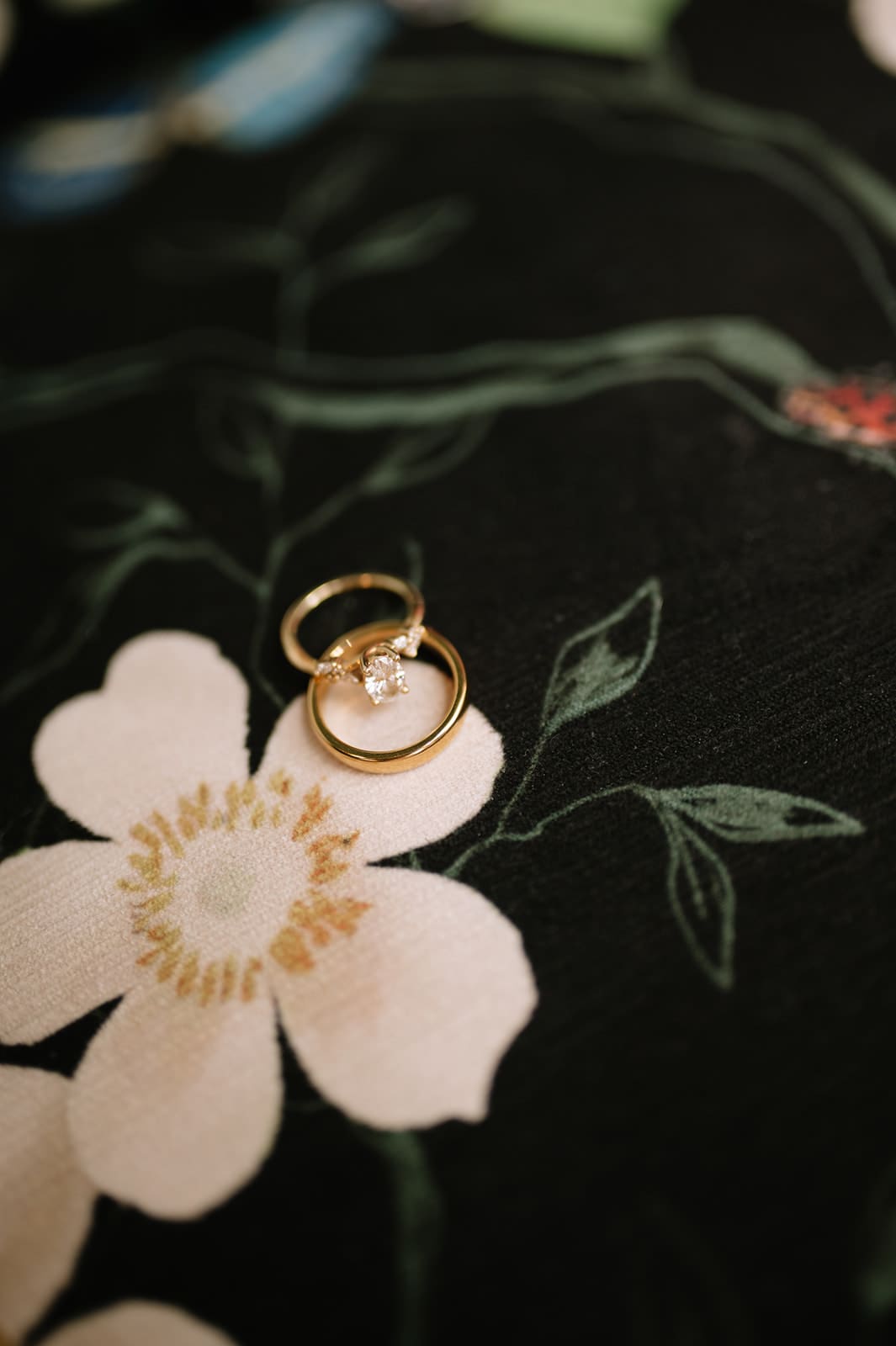 Two gold rings, one with a small diamond, rest on a dark floral-patterned fabric with white flowers and green leaves.