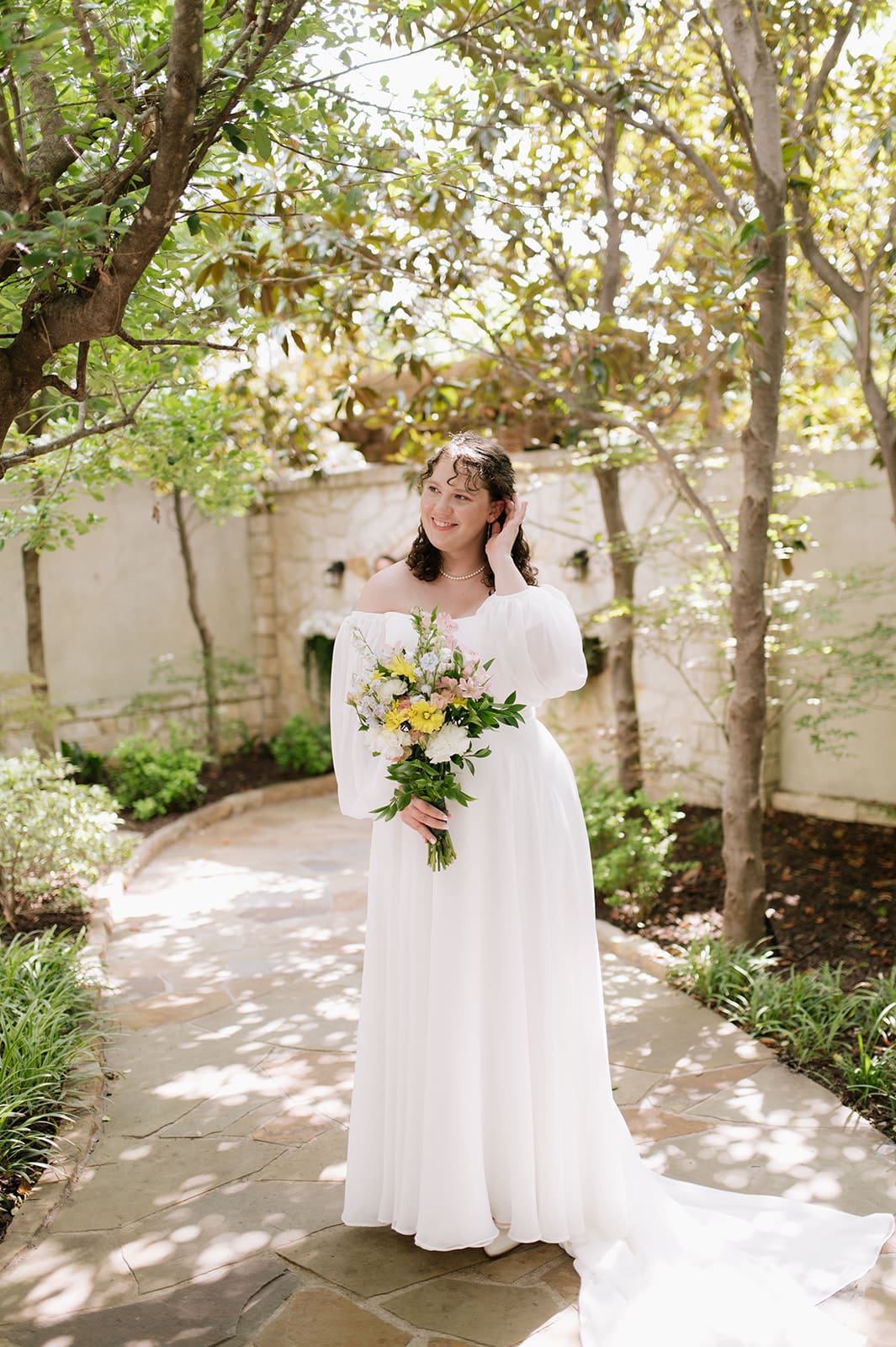 A group of six people, including a bride in a white dress and groom in a green suit, pose together outdoors, smiling at the camera at a wedding at Magnolia Terrace in Frisco, Tx