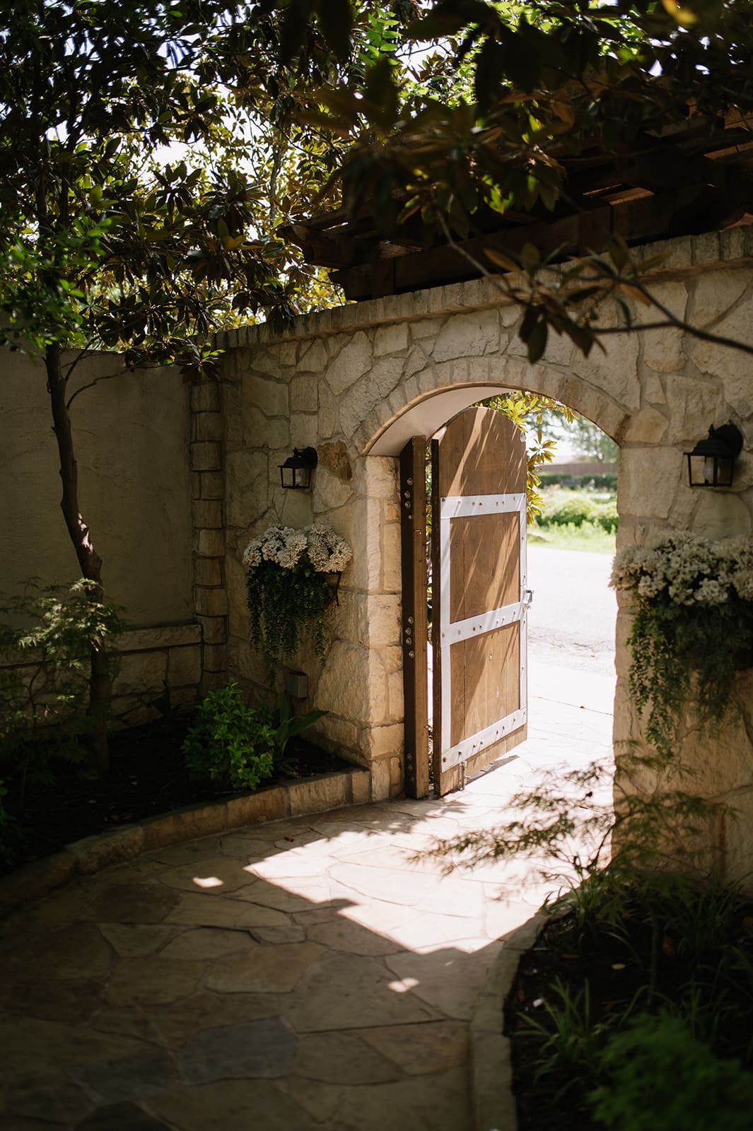 A wooden gate set in a stone wall stands open, letting sunlight stream onto a stone pathway surrounded by greenery and potted flowers.