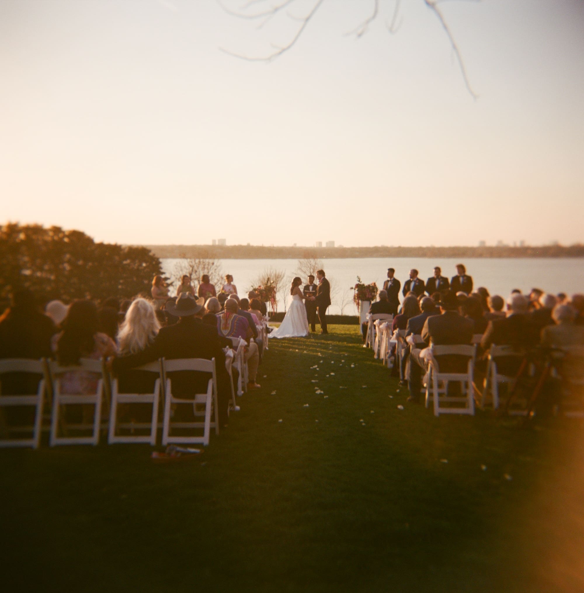Outdoor wedding ceremony by a lake at sunset, with a couple at the altar, wedding party standing on each side, and guests seated in rows for a  Dallas Arboretum Wedding