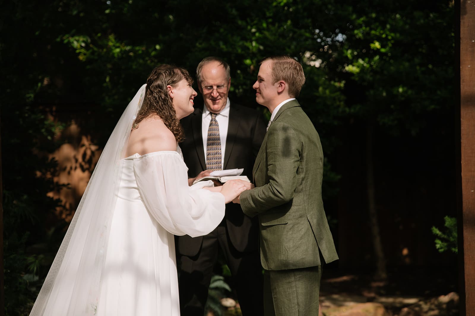 A bride and groom stand facing each other, holding hands and smiling, with an officiant standing behind them during an outdoor wedding ceremony at Magnolia Terrace in Frisco, Tx