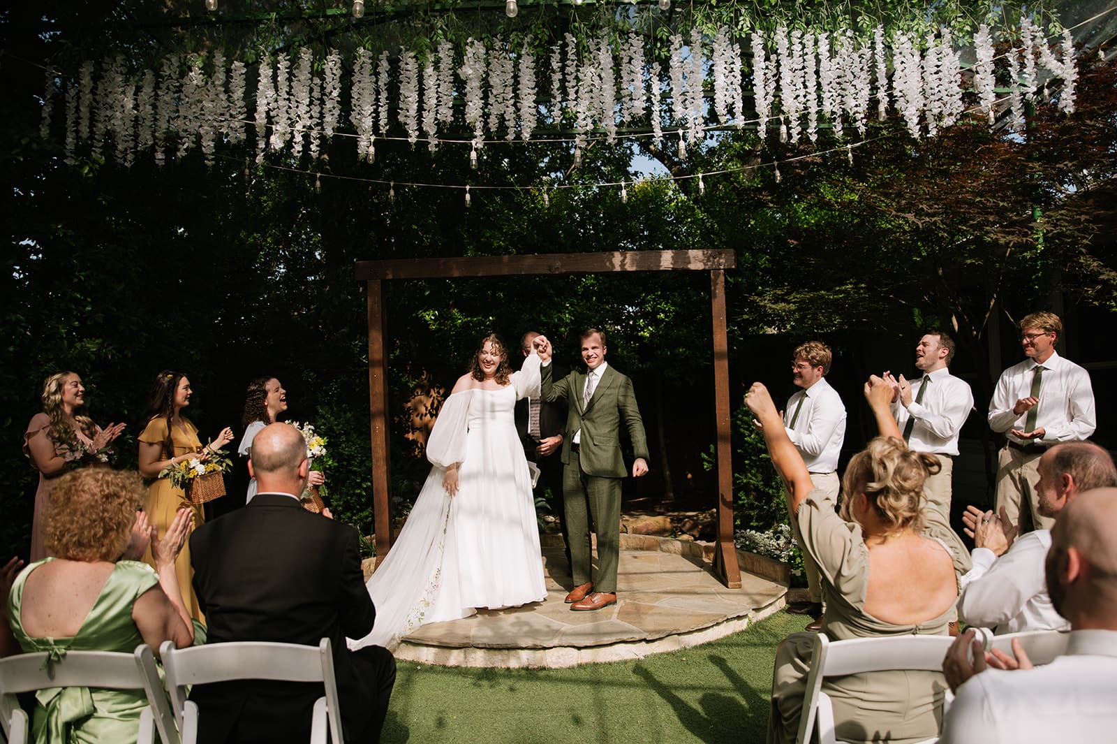 A bride and groom stand under an outdoor arch holding hands as seated guests and attendants applaud during a daytime wedding ceremony.