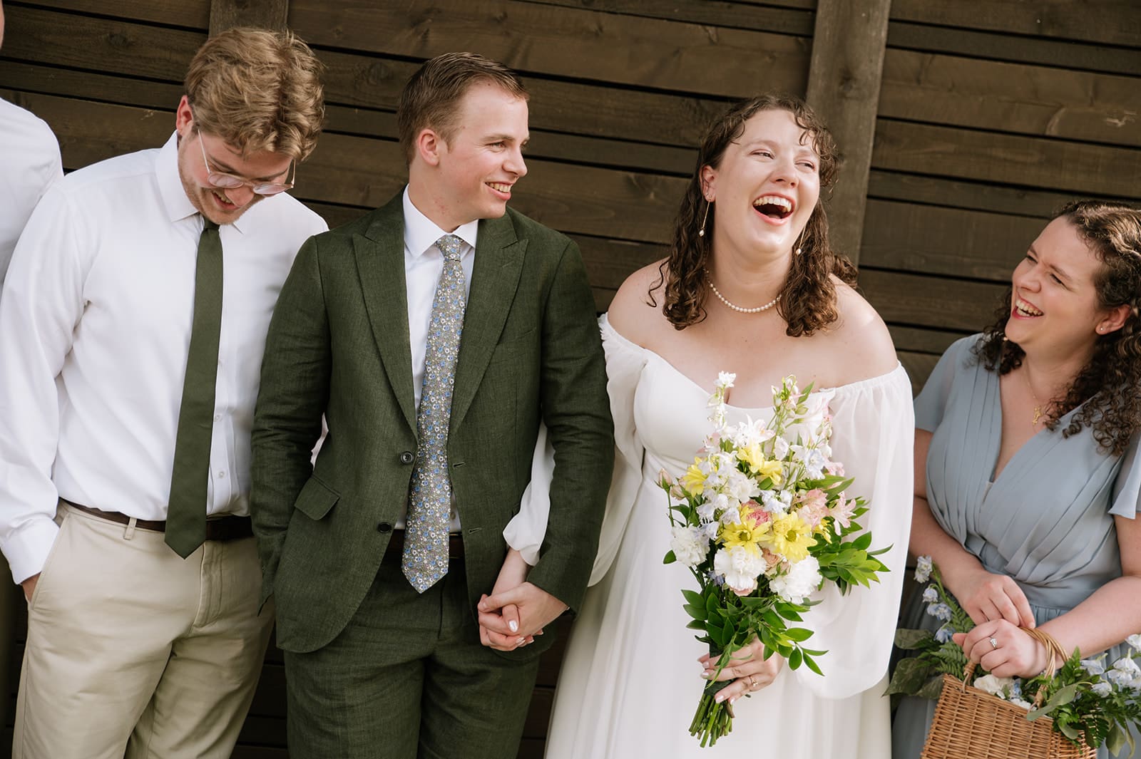 A group of six people, including a bride in a white dress and groom in a green suit, pose together outdoors, smiling at the camera at a wedding at Magnolia Terrace in Frisco, Tx