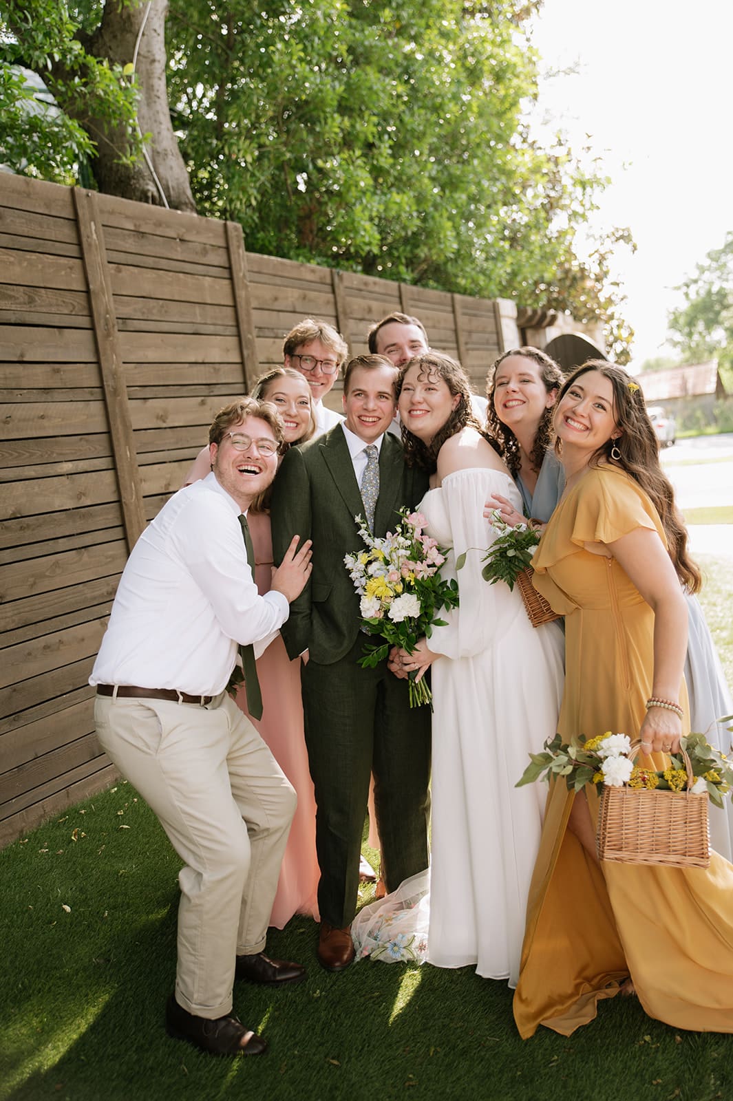 A group of six people, including a bride in a white dress and groom in a green suit, pose together outdoors, smiling at the camera at a wedding at Magnolia Terrace in Frisco, Tx