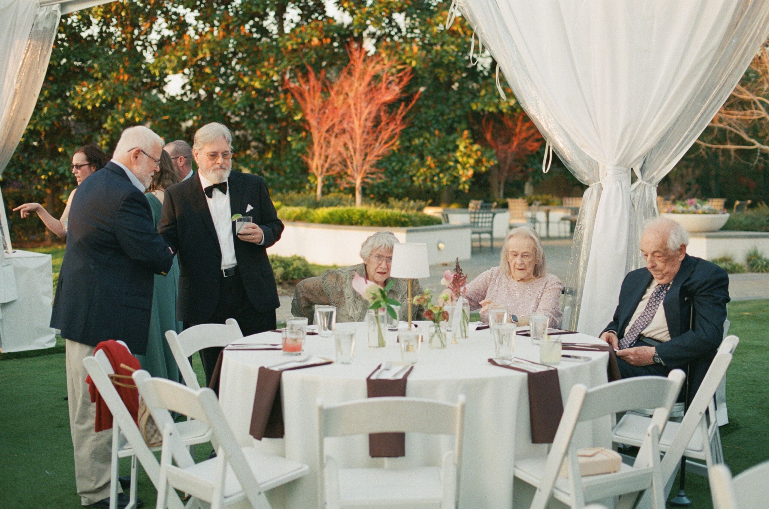 Long table set for an event under a clear tent with string lights, surrounded by guests and white chairs at the Dallas Arboretum Wedding