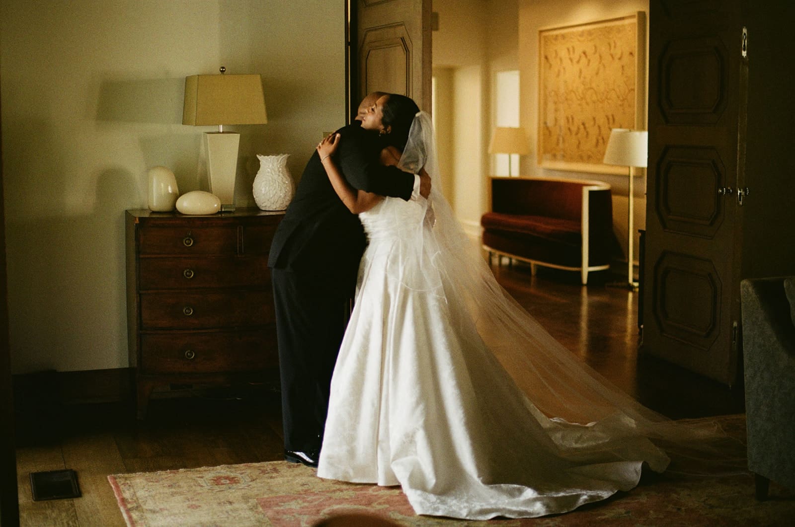 A bride in a white wedding dress and veil hugs a person in a suit inside a softly lit, elegant room for a Dallas Arboretum Wedding