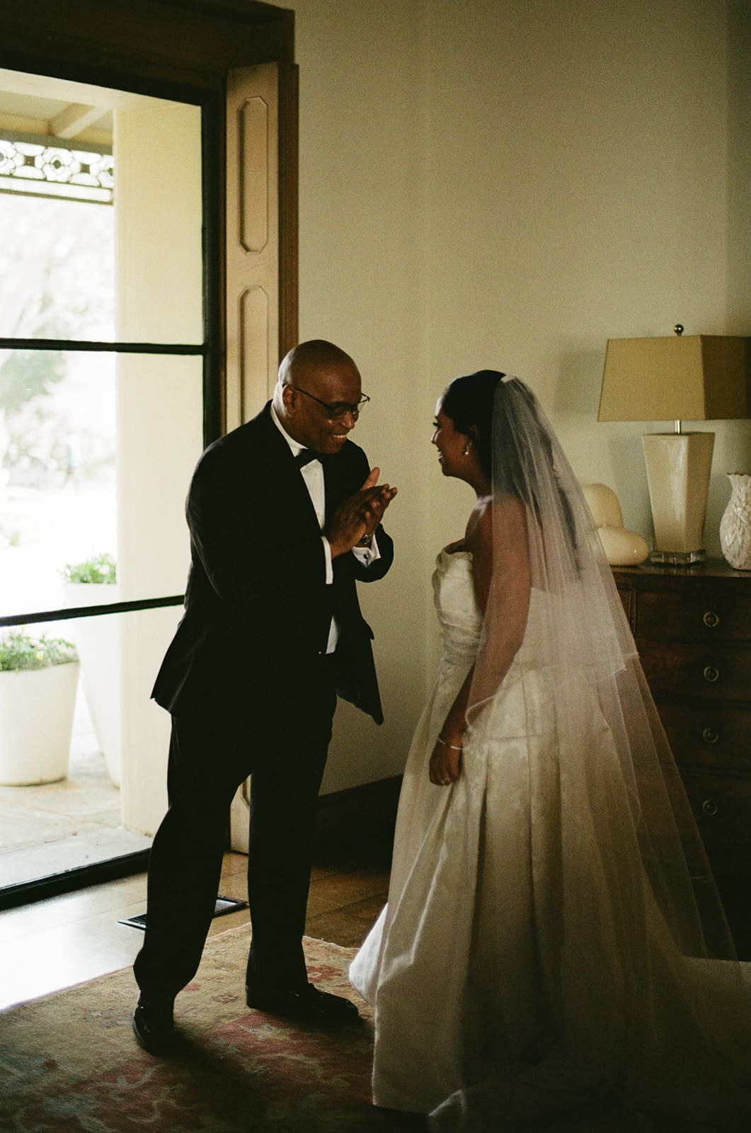 A man in a suit and a woman in a wedding dress stand indoors near a doorway, engaged in conversation.