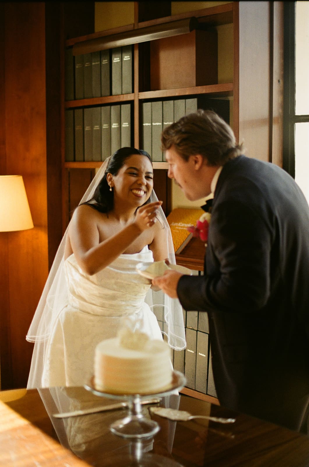 A bride and groom, dressed in wedding attire, smile and feed each other cake in a room with bookshelves and a lamp.