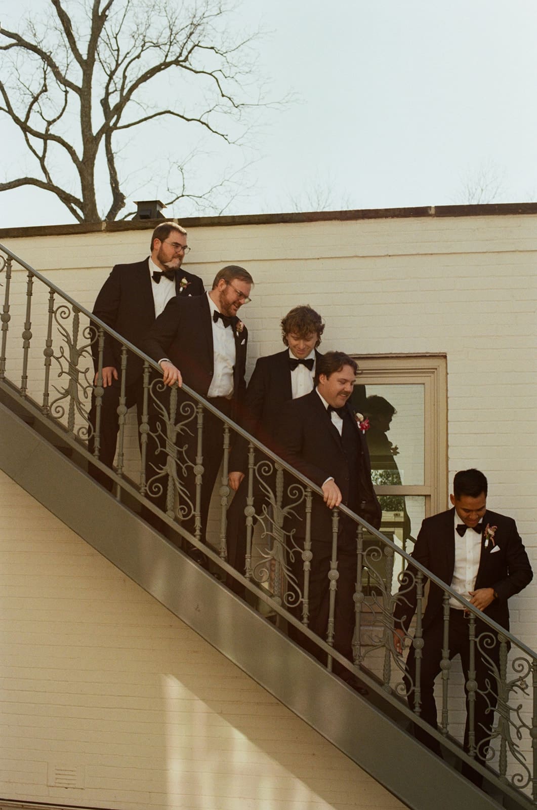 Five men in tuxedos stand and walk down an outdoor staircase next to a building with a window, with bare tree branches visible in the background for a Dallas Arboretum Wedding