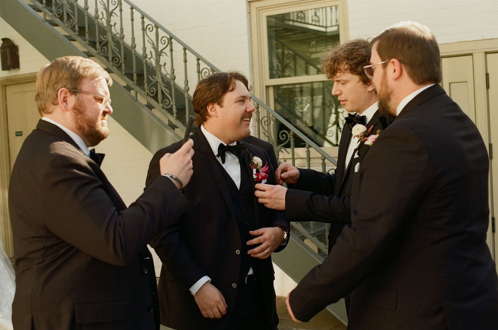 Three men in black suits stand near a building with green shutters; one adjusts his jacket, another gestures with his hand, and a third stands smiling in the background.