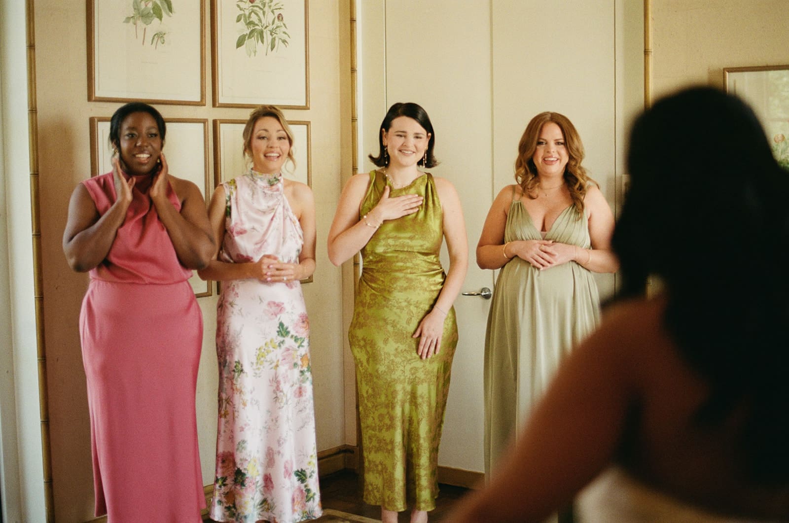 A bride in a white dress stands with four women in colorful dresses in a room with floral artwork on the wall.