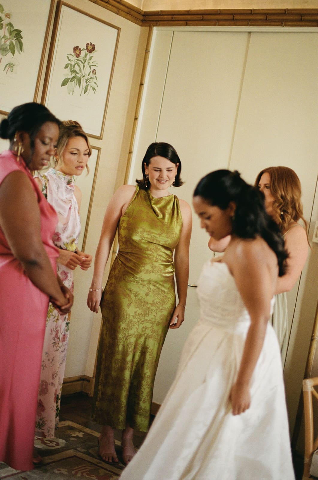 A bride in a white dress stands with four women in colorful dresses in a room with floral artwork on the wall.