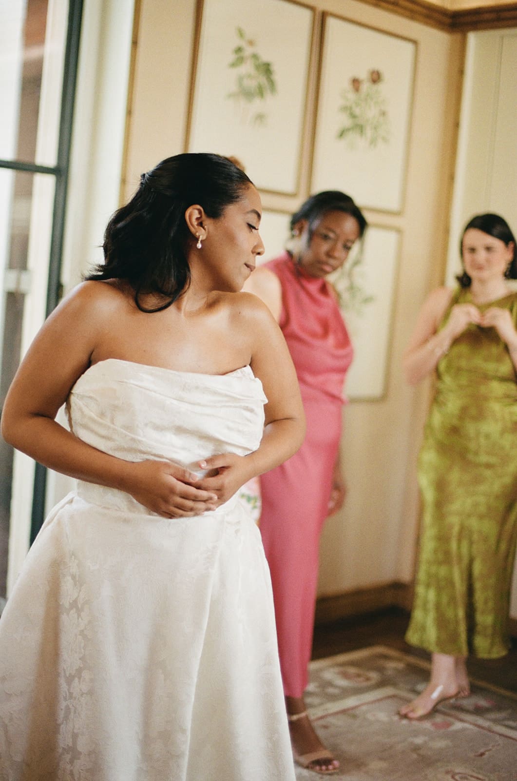A bride in a white dress stands with four women in colorful dresses in a room with floral artwork on the wall.