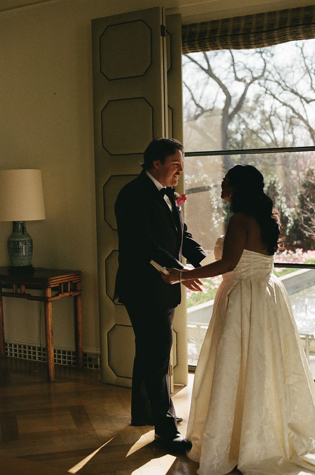 A bride and groom stand facing each other indoors near a large window, holding hands and talking at a  Dallas Arboretum Wedding
