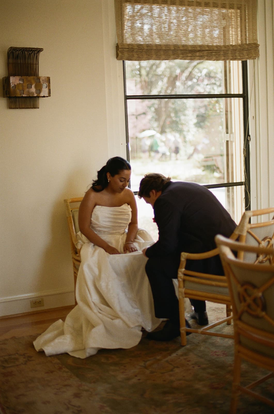 A bride in a white dress sits on a chair while a person in a suit kneels in front of her, adjusting or helping with her shoe near a window at a Dallas Arboretum Wedding