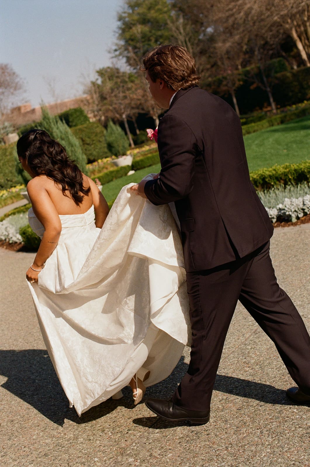 A bride and groom walk outdoors; the groom holds up part of the bride’s white dress as they move along a sunlit path in a garden setting.