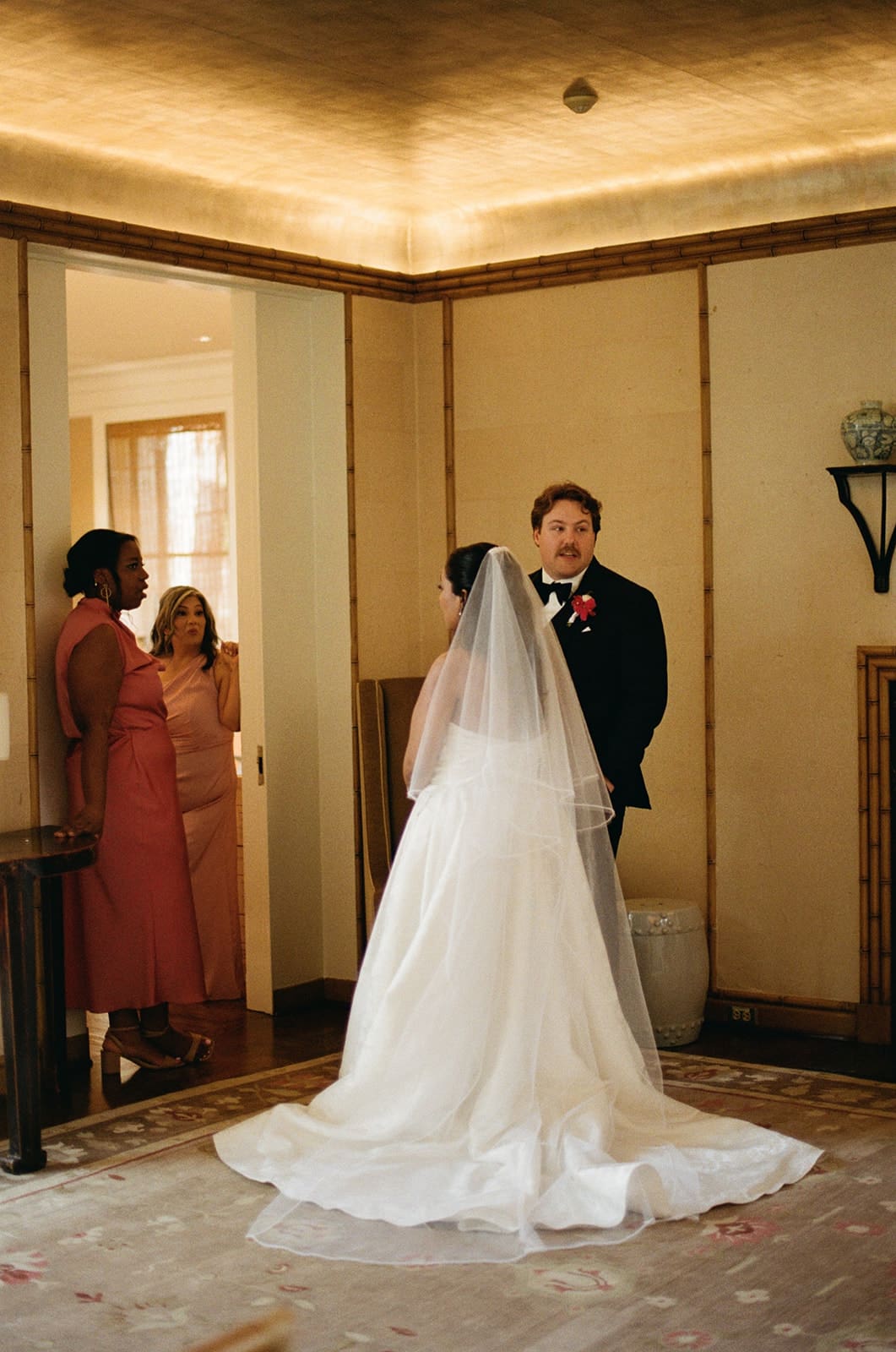 A bride in a white gown and veil stands facing a groom in a suit, with two women in pink dresses watching from a doorway in a warmly lit room.