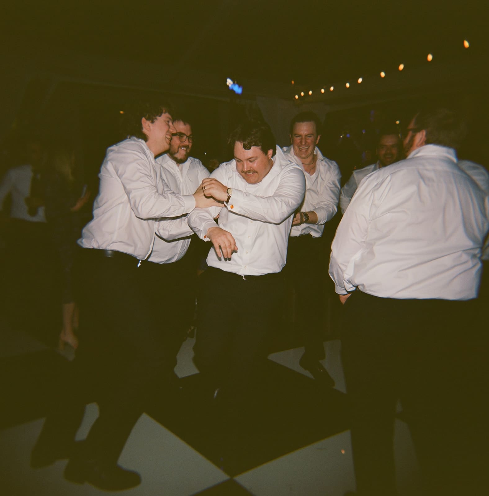 A group of men in white shirts and dark pants are dancing and laughing together on a black-and-white checkered floor.