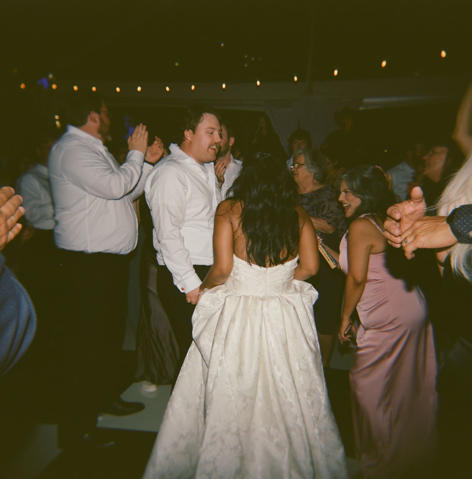 A bride in a white dress and guests in formal attire dance together at a lively indoor celebration for a Dallas Arboretum Wedding