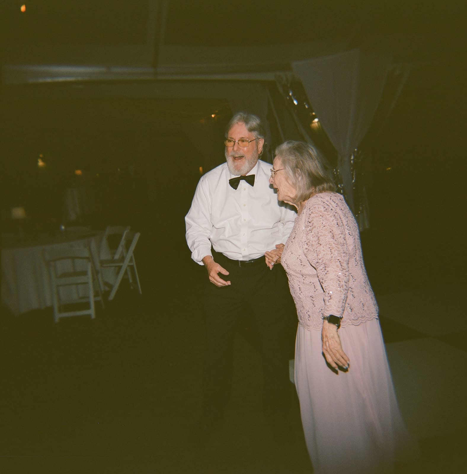 An older man in a white shirt and bow tie stands next to an older woman in a pink dress. They are both smiling in a dimly lit room.