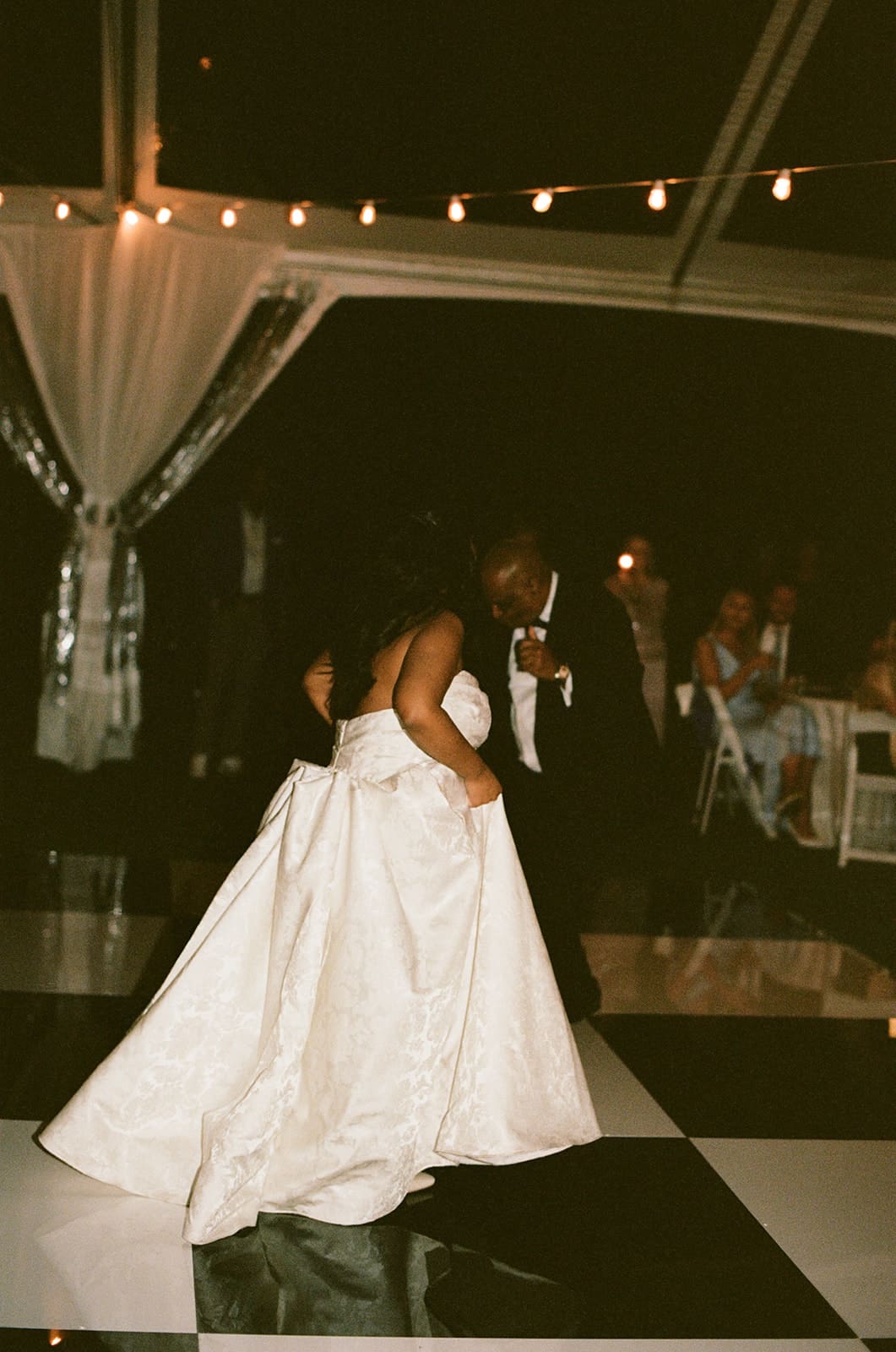 A bride in a white dress and guests in formal attire dance together at a lively indoor celebration for a Dallas Arboretum Wedding