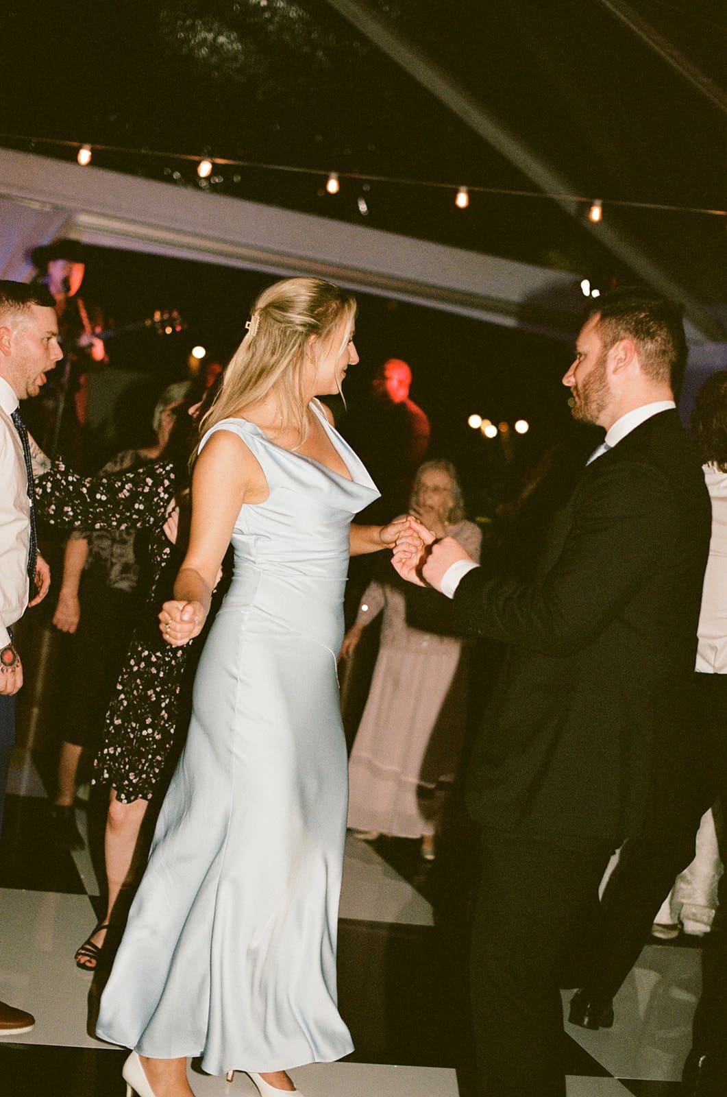 A bride in a white dress and guests in formal attire dance together at a lively indoor celebration for a Dallas Arboretum Wedding