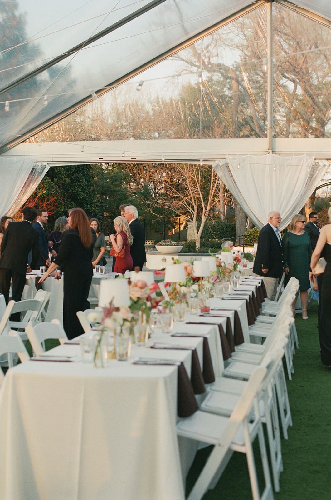 A long table set for an outdoor event under a clear tent, with guests standing and mingling in the background for a Dallas Arboretum Wedding