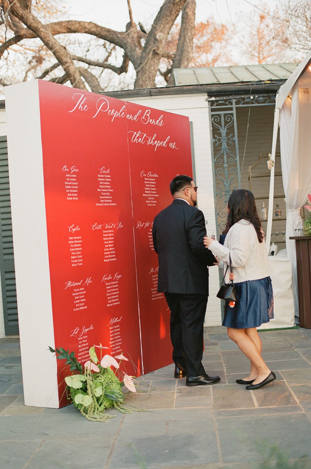 Two people stand in front of a large red seating chart displayed outdoors at an event, reading the names and table assignments listed on it.
