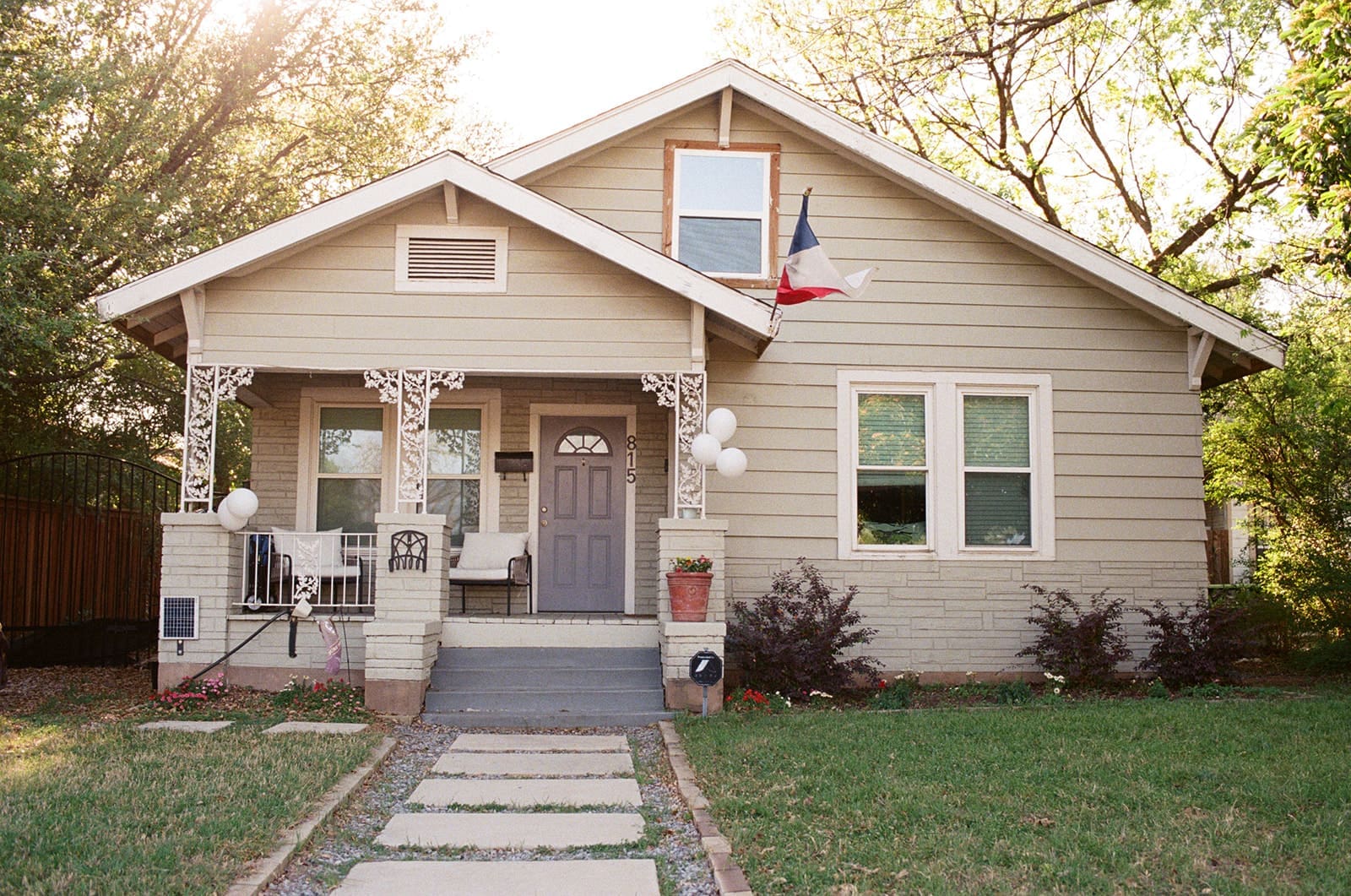 Single-story beige house with a front porch, chairs, white balloons, and a Texas flag, surrounded by grass and trees on a sunny day.