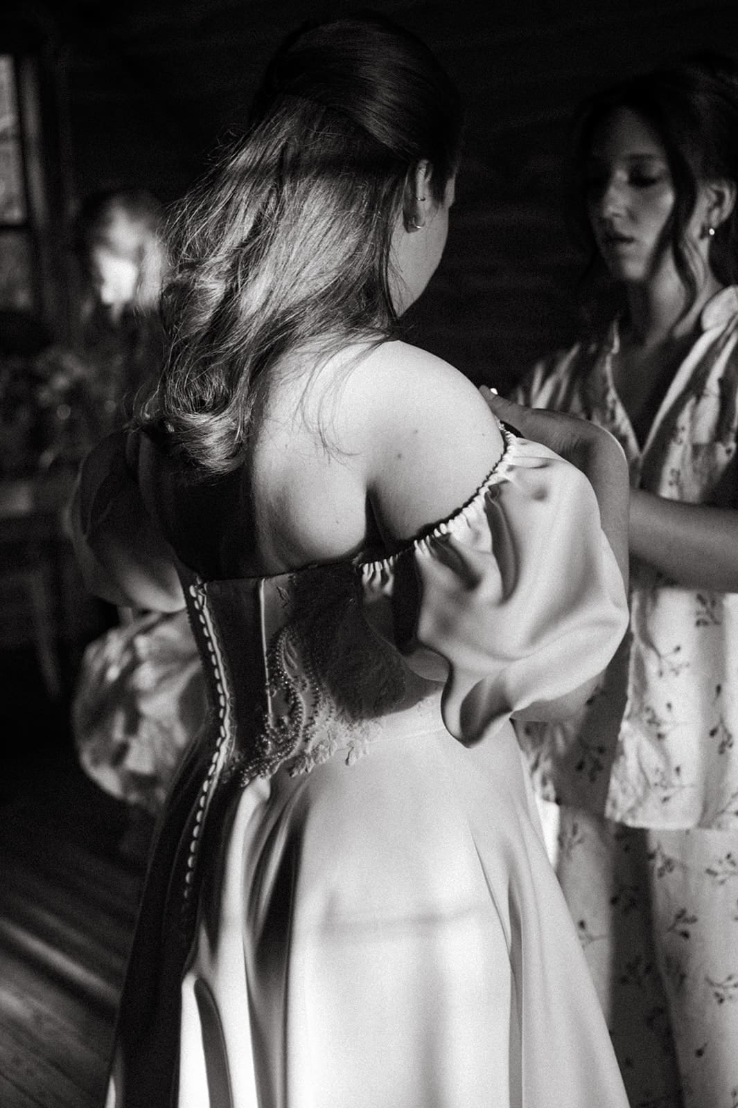 A woman in a dress is being assisted by another person in a softly lit room; the image is in black and white.