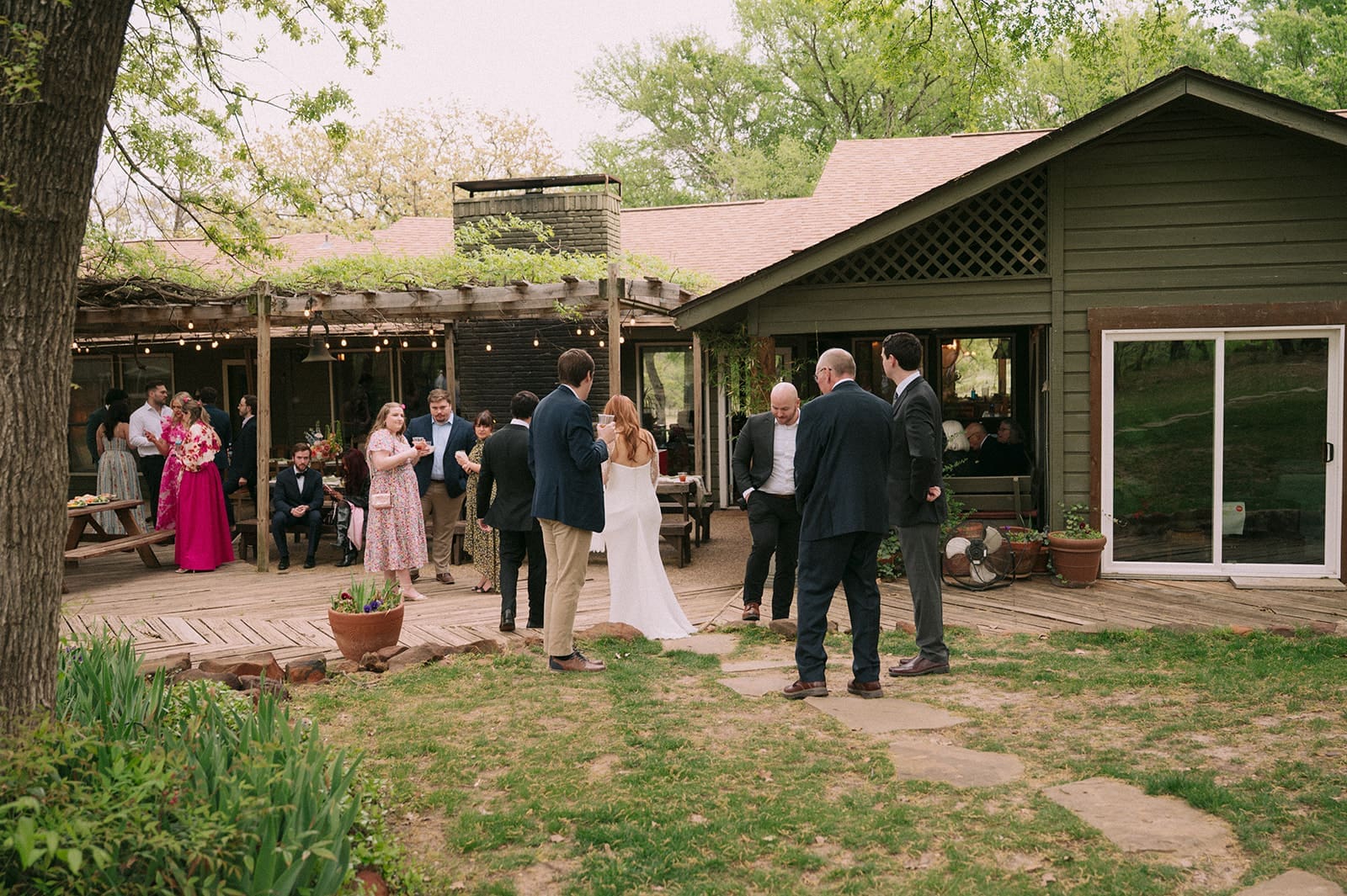 A small group of people, some dressed formally, gather outside a house with a patio during a daytime outdoor event.