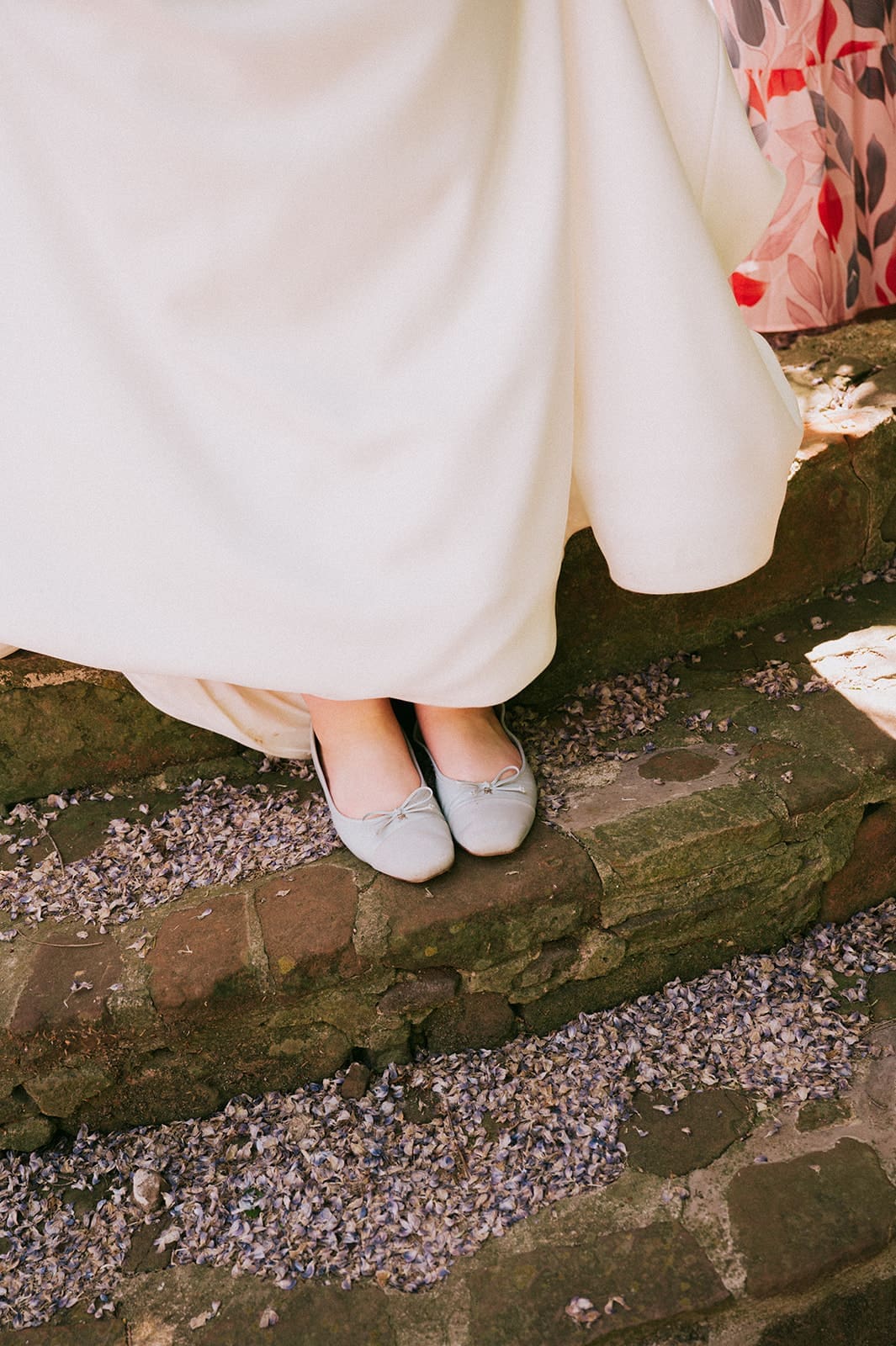 A person in a white dress stands on stone steps, showing only their lower legs and light-colored shoes.