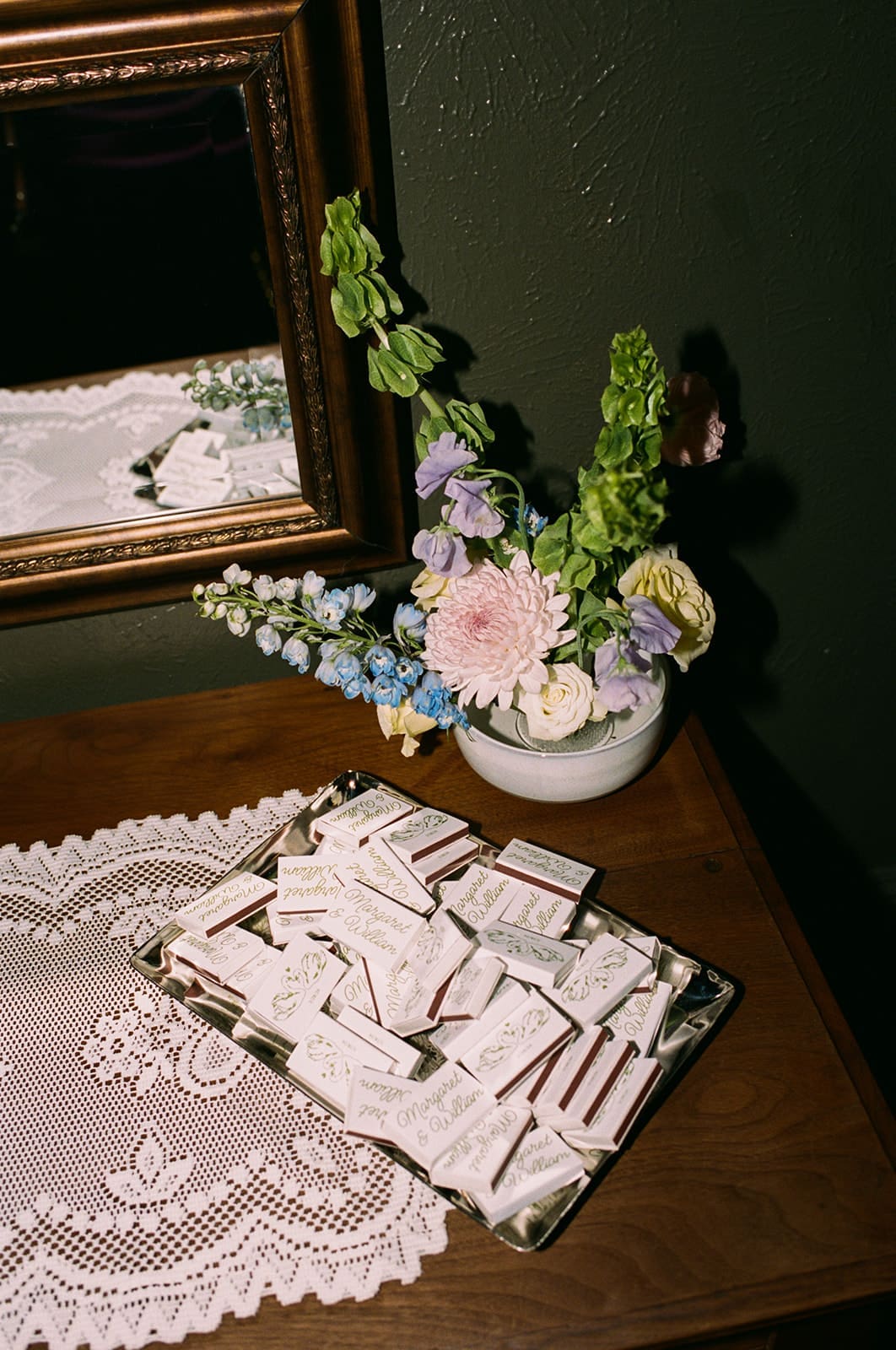 A tray with name tags sits on a wooden table next to a lace doily and a vase of mixed flowers.