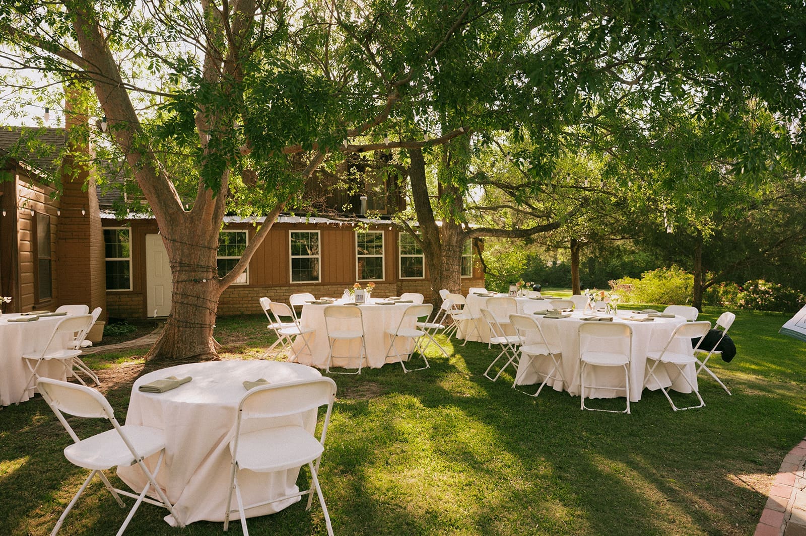 Round tables with white tablecloths and folding chairs set up outdoors near a brown wooden building, prepared for an event for a weston gardens wedding