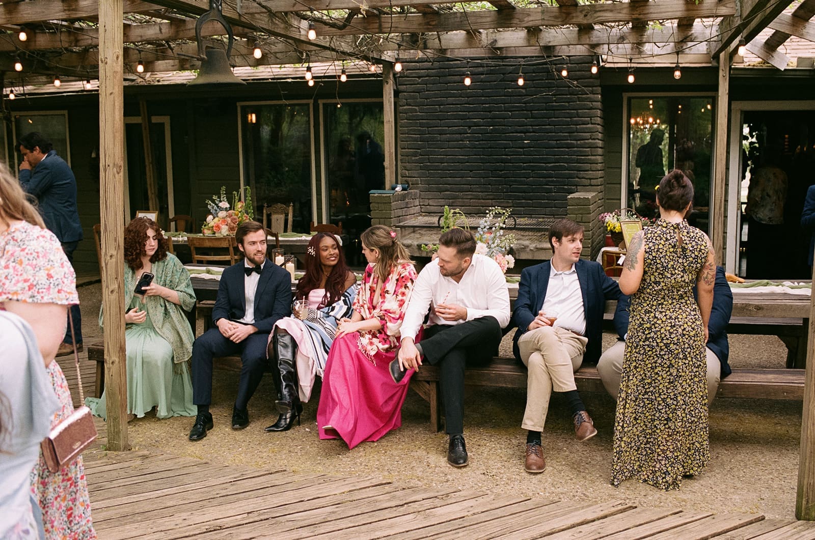 A small group of people, some dressed formally, gather outside a house with a patio during a daytime outdoor event.