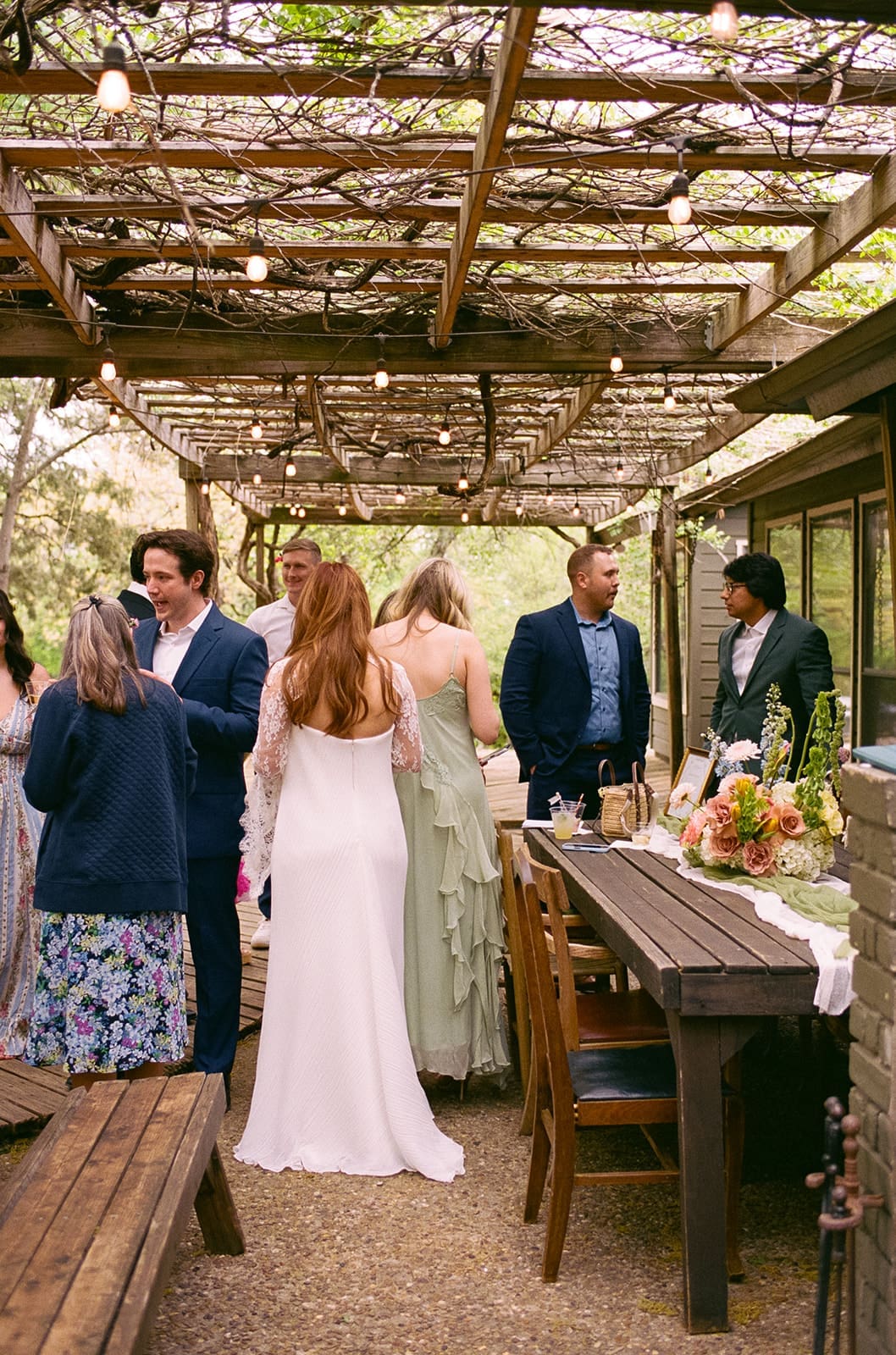 A group of people in formal attire gather and converse around a long outdoor dining table under a pergola decorated with string lights.