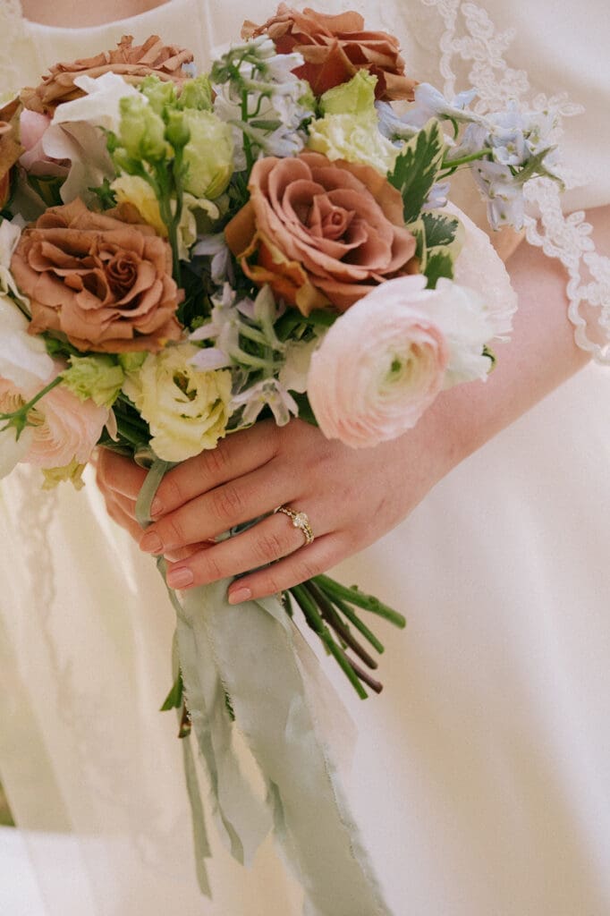 A person in a white lace dress holds a bouquet of brown, pink, and white flowers, with a ring visible on their finger.